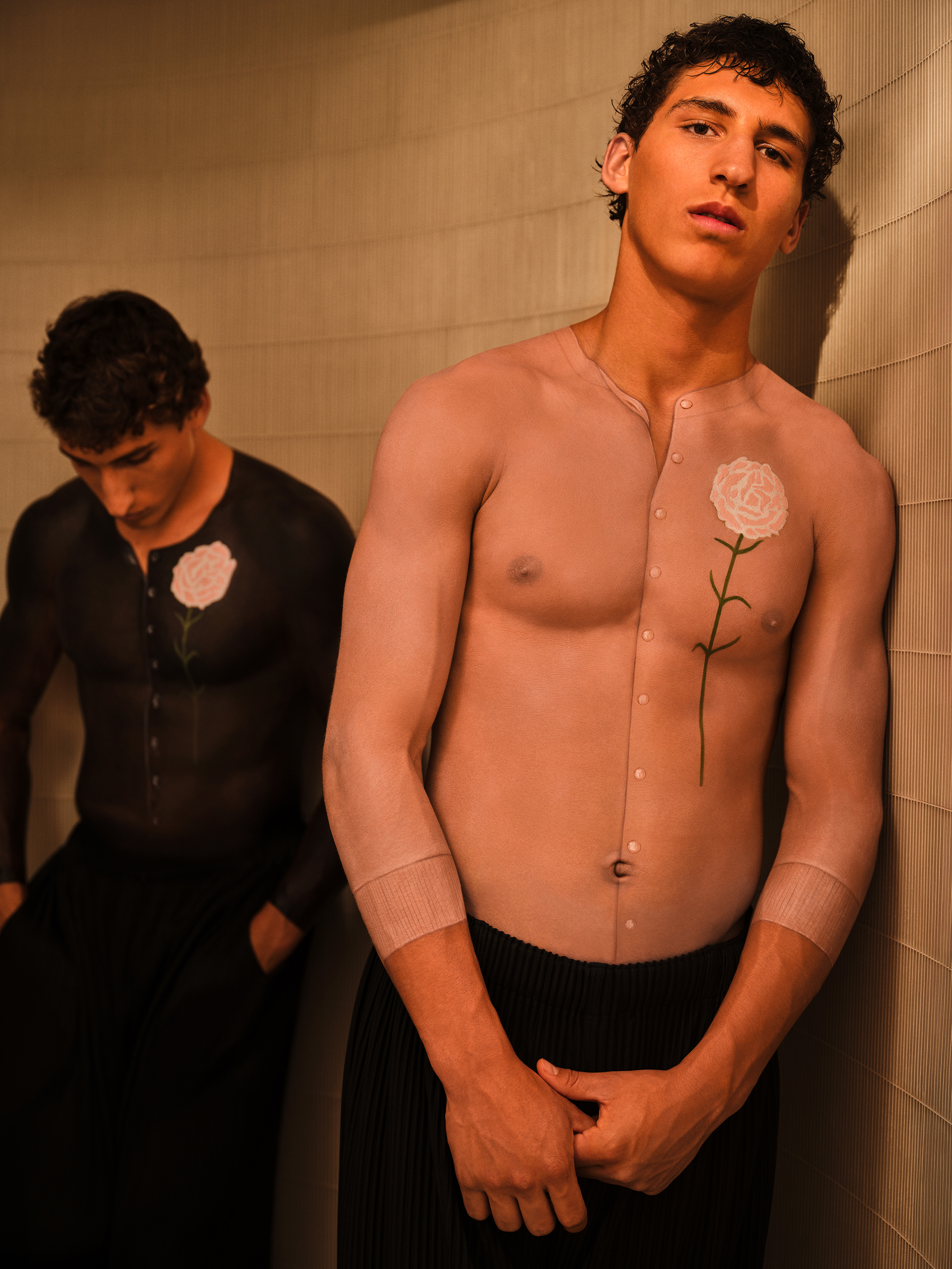 Twin brothers leaning against a grey wall, each with body paint on their chests which makes it appear that they are wearing button up sweaters